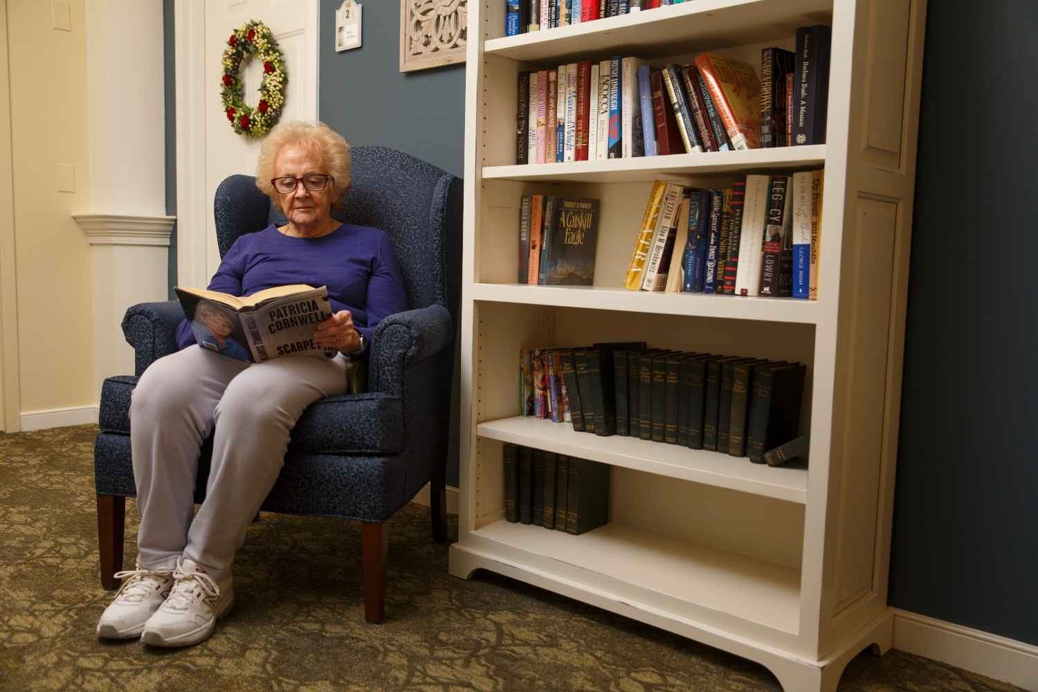 An elderly woman sits in a comfortable armchair reading a book next to a tall bookshelf.