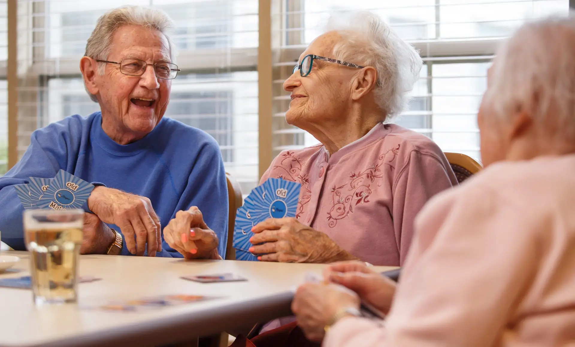 Three joyful seniors playing Old Maid card game at a table with drinks.