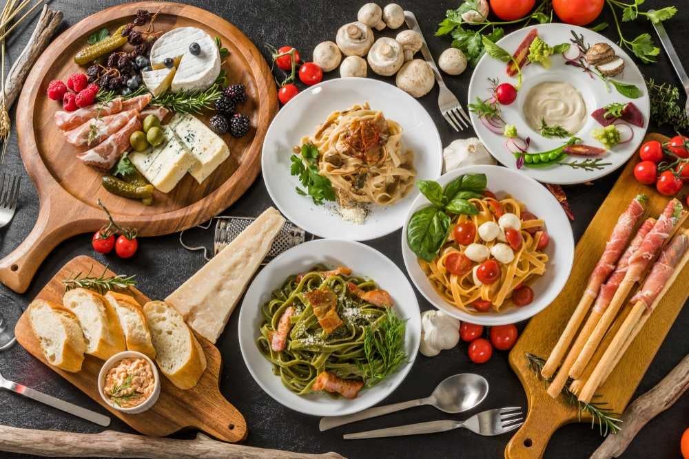 A feast of Italian dishes: antipasto with cheeses and meats, three pasta plates, and breadsticks with prosciutto.