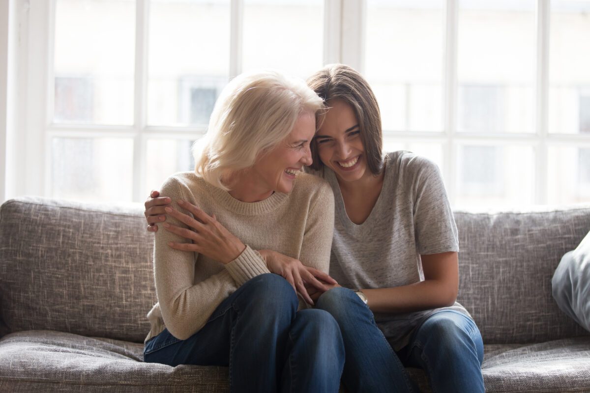 A woman and her senior mother sit on a couch, laughing together and embracing.