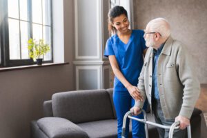 An elderly man with a walker is escorted by a smiling healthcare worker wearing blue scrubs.