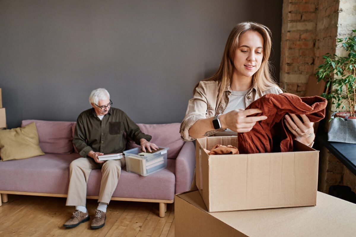 An adult child helps pack up their older parent’s belongings while the parents sit in the background sorting a box.