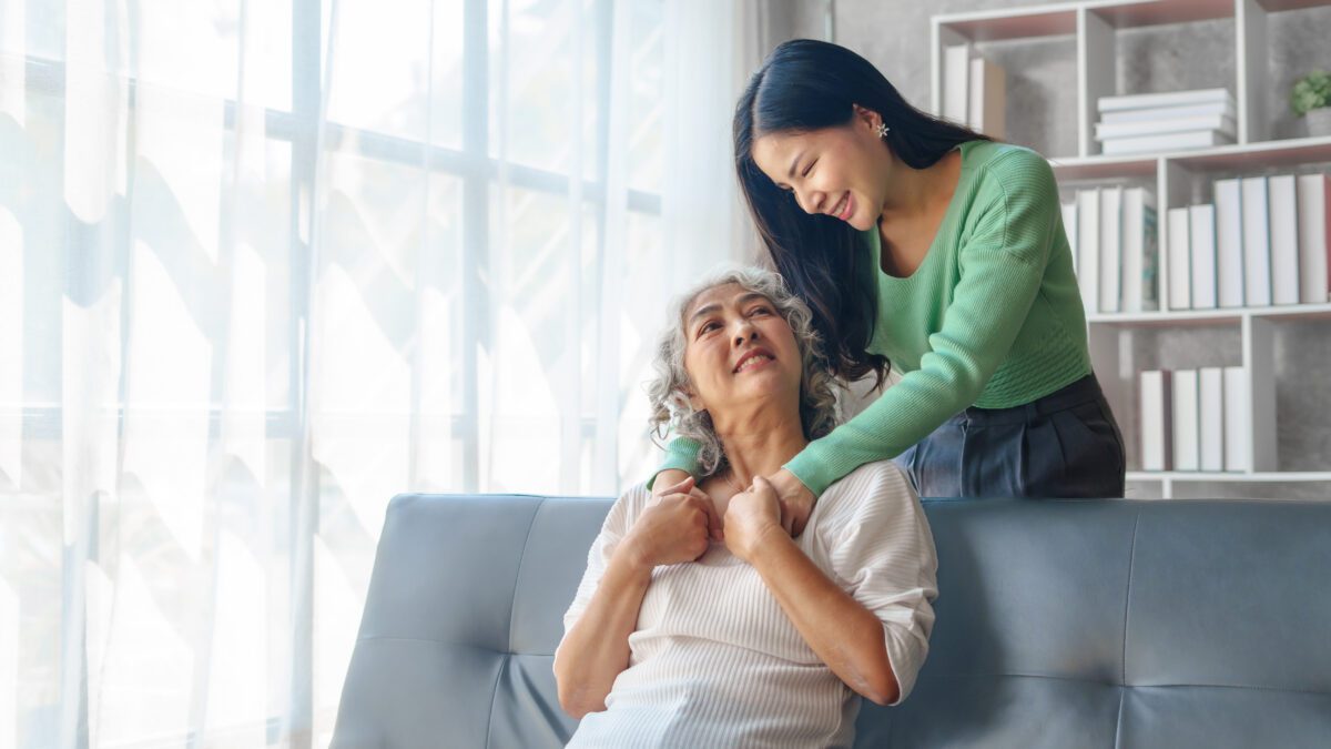 A younger woman with dark hair and a green shirt embraces her mother with gray hair and a white shirt over the back of a couch.