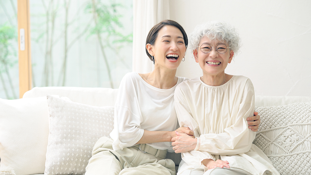 A woman sits with her elderly parent, and they are both smiling widely.