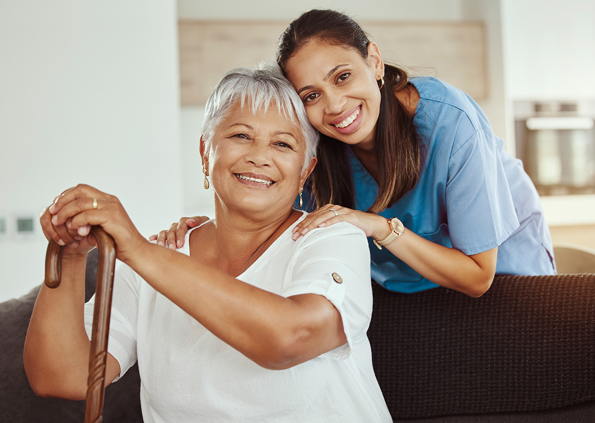 Two women with touching heads, one the caregiver dressed in blue and one the resident dressed in white, smile at the camera.