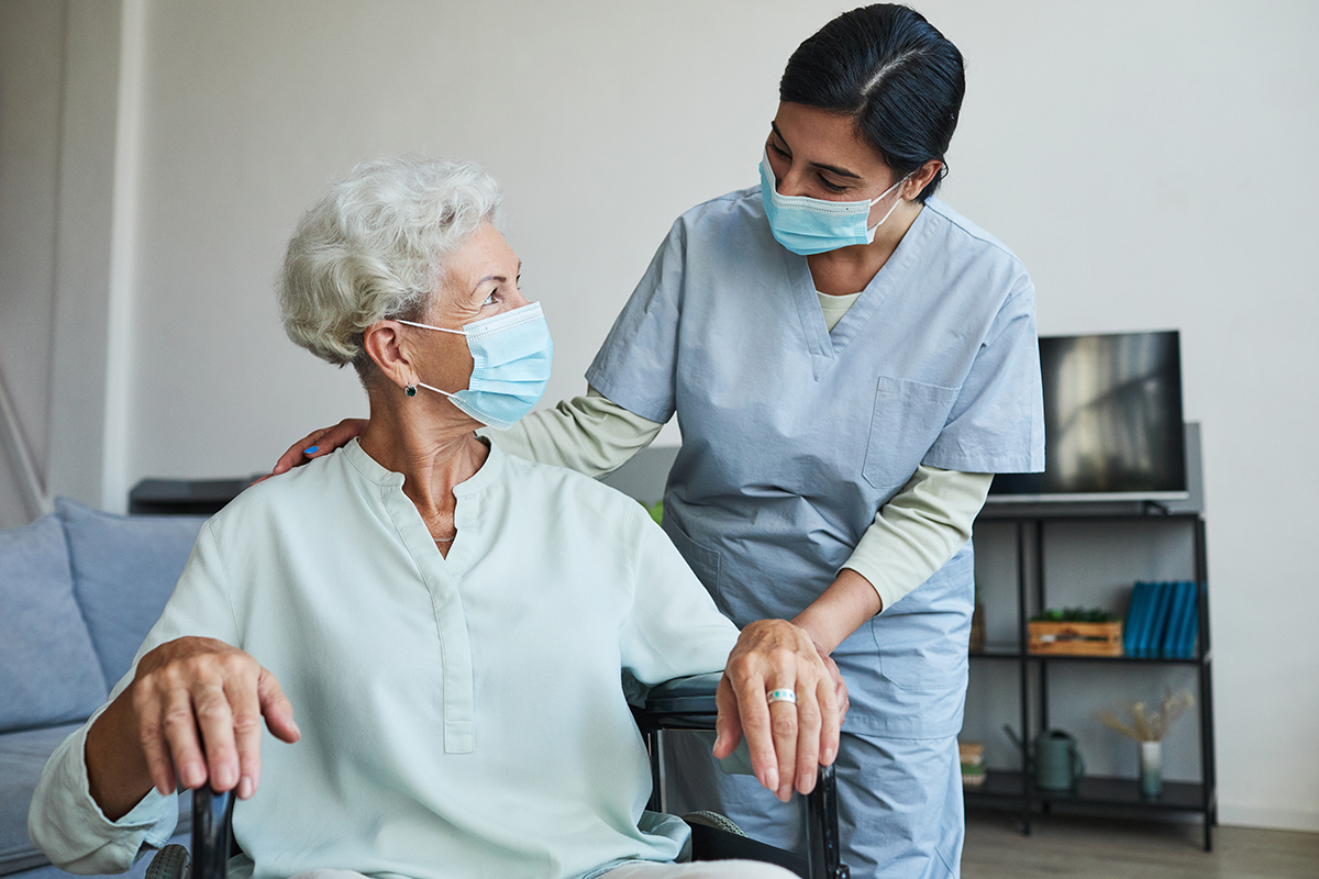 An image of a woman in a wheelchair and a medical professional facing each other, both wearing masks.