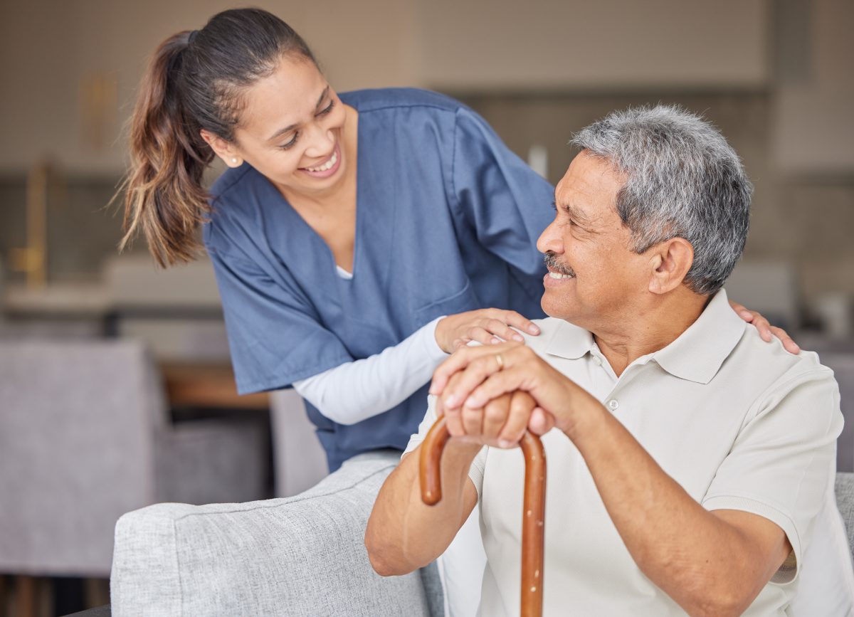 a healthcare provider smiling while leaning over the shoulder of a smiling elderly man who has a cane.