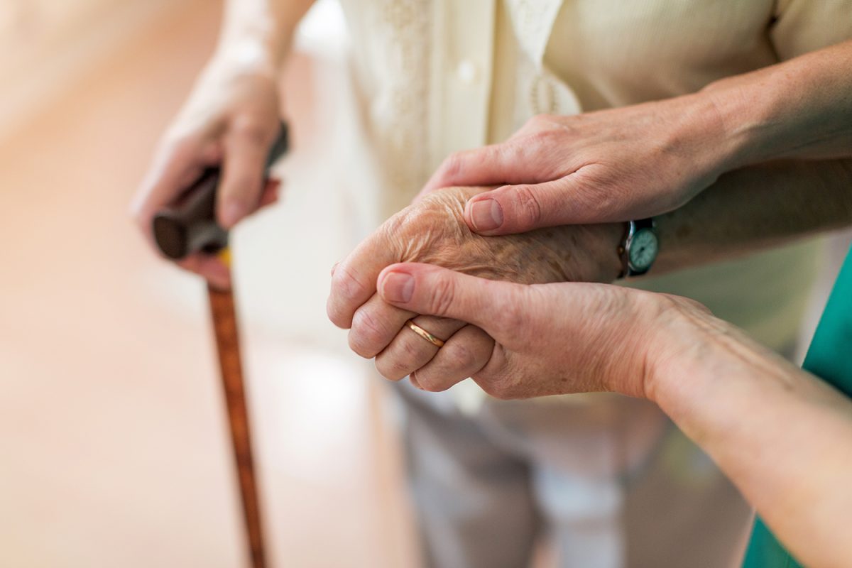 A person holds the hand of an older person who is wearing a gold ring and a watch, and holding a cane.